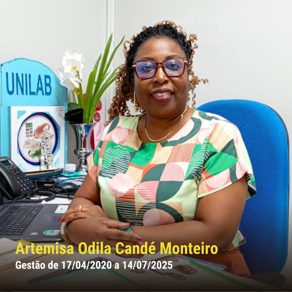 Mulher negra de pele escura, com óculos e blusa colorida geométrica, sentada sorrindo em mesa com materiais da UNILAB.