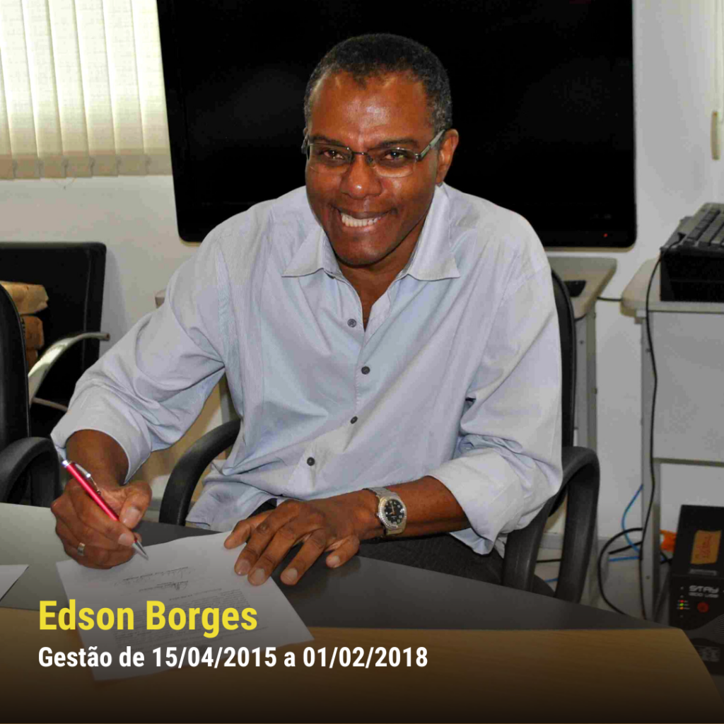 Homem negro de pele escura, com óculos e camisa clara, sorrindo enquanto assina documento em ambiente institucional.