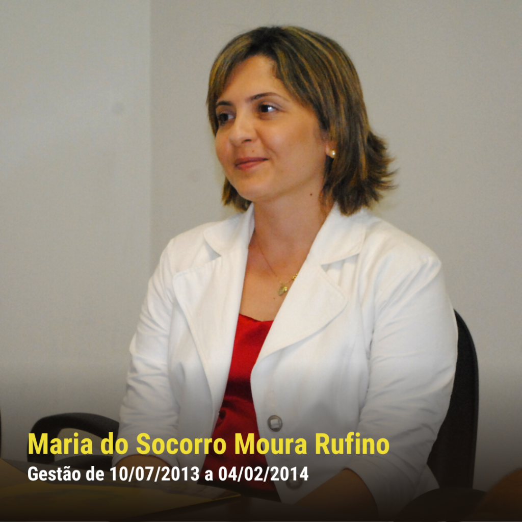 Mulher branca de pele clara, com blazer branco e blusa vermelha, sentada em sala de reunião, olhando levemente para o lado.
