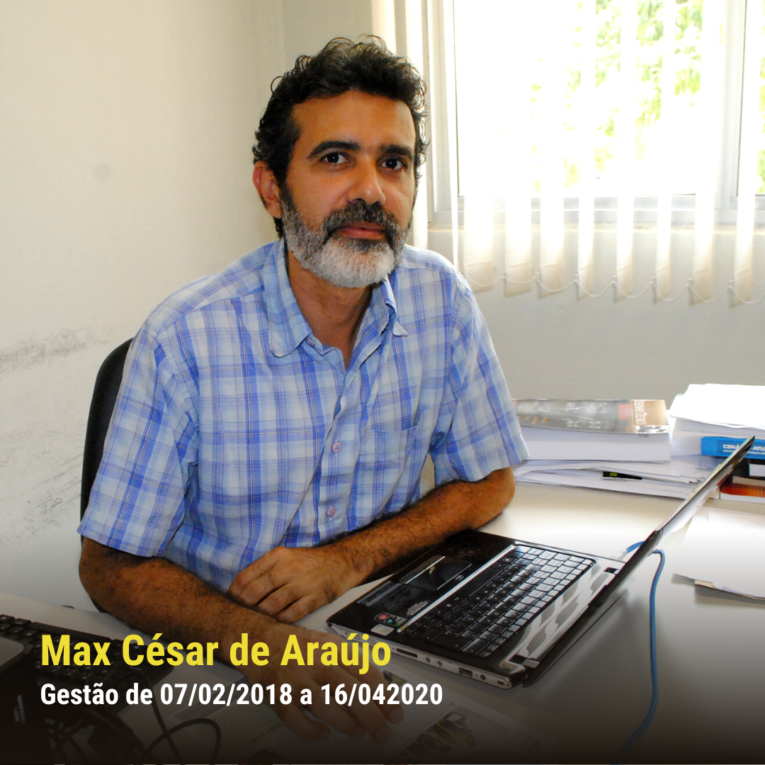 Homem pardo de pele clara, com barba e camisa xadrez azul, sentado à mesa com notebook e livros, olhando para a câmera.