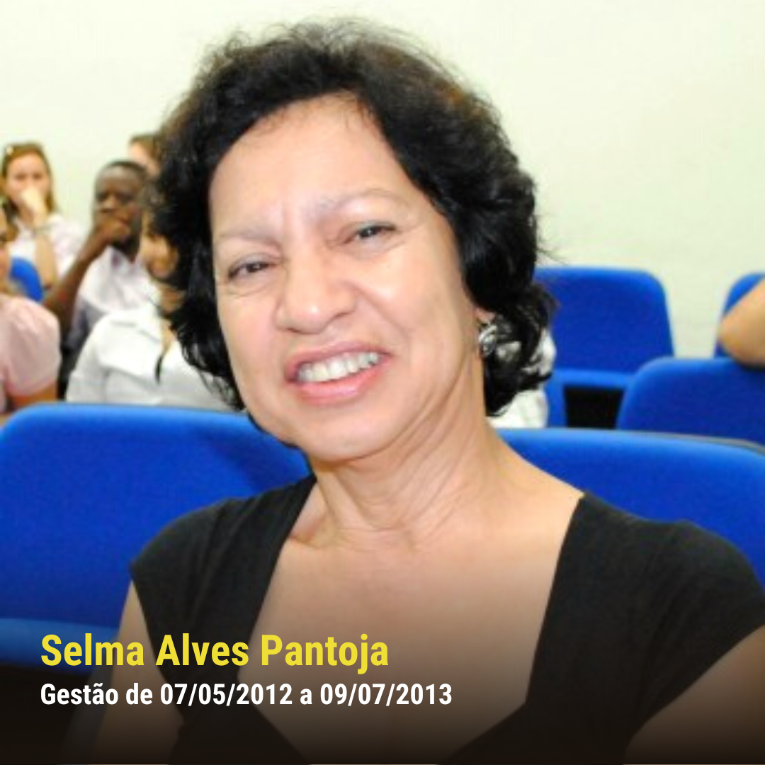 Mulher branca de pele clara, com cabelo curto e blusa preta, sorrindo sentada em auditório com cadeiras azuis.
